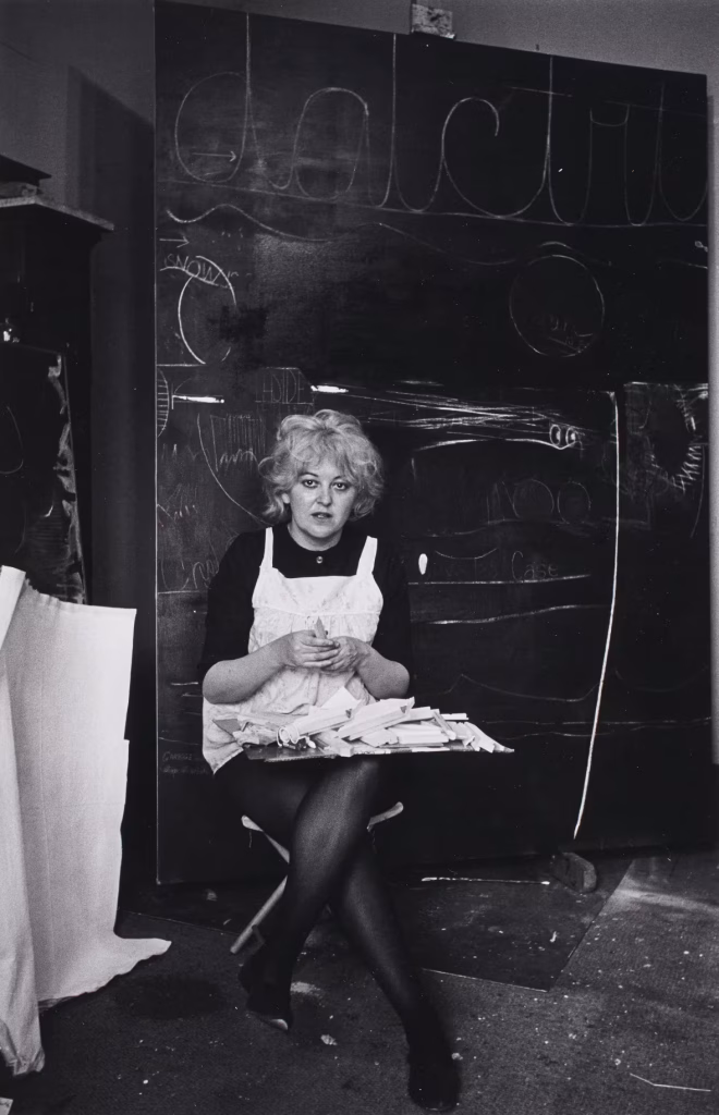 JA photograph of the artist Joyce Wieland sitting with handiwork against a black background wearing black clothing and a white apron